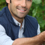 Smiling man wearing a navy vest and checkered shirt outdoors.