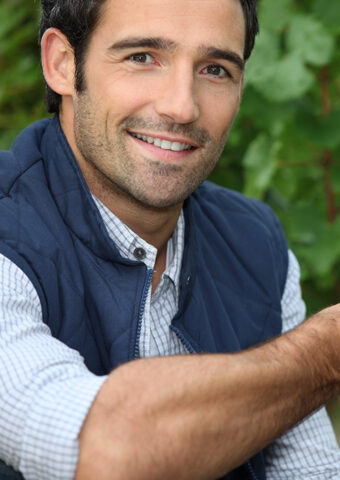 Smiling man wearing a navy vest and checkered shirt outdoors.